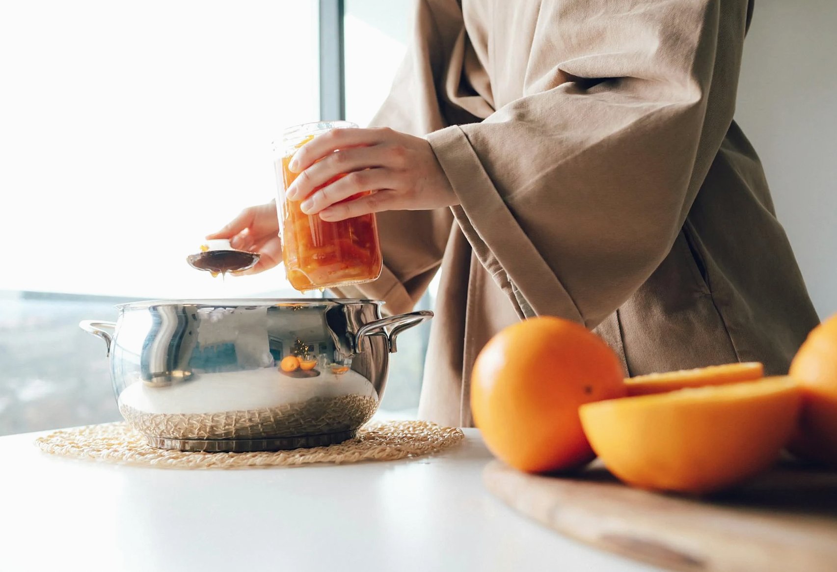 Una persona añade miel a una olla junto a naranjas frescas sobre una mesa, en un entorno de cocina.